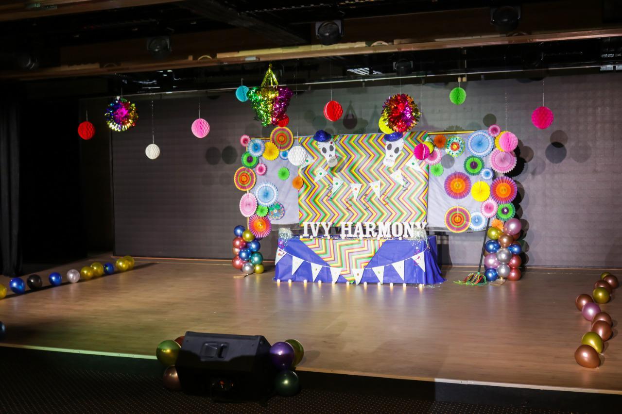 The image showcases a vibrant and festive stage setup at IVY STEM International School. The backdrop features a colorful geometric pattern with the words 